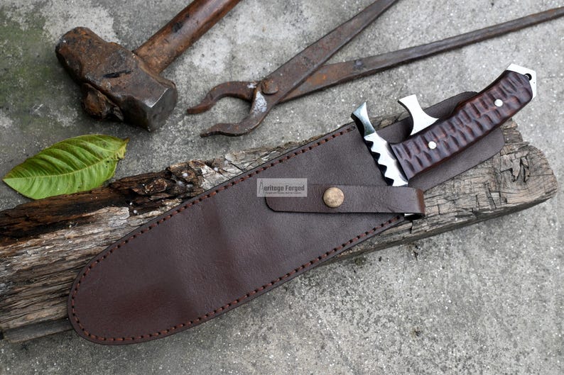 Puede incluir: Una funda de cuero marr&oacute;n con un borde cosido y un bot&oacute;n de metal. Un cuchillo con un mango de madera marr&oacute;n y una hoja plateada est&aacute; dentro de la funda. La funda est&aacute; sobre un trozo de madera.
