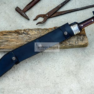 May include: A black leather scabbard with a silver buckle and a brown wooden handle knife with a silver metal guard. The knife is on a piece of wood with the text "Heritage Forged" on it.