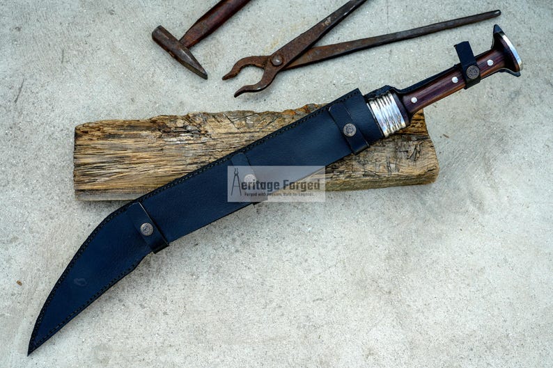 May include: A black leather scabbard with a silver buckle and a brown wooden handle knife with a silver metal guard. The knife is on a piece of wood with the text "Heritage Forged" on it.