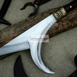 May include: A silver-colored curved blade with a wooden handle and a brass decorative piece. The blade is on a wooden surface. The text "Heritage Forged" is visible on the blade.