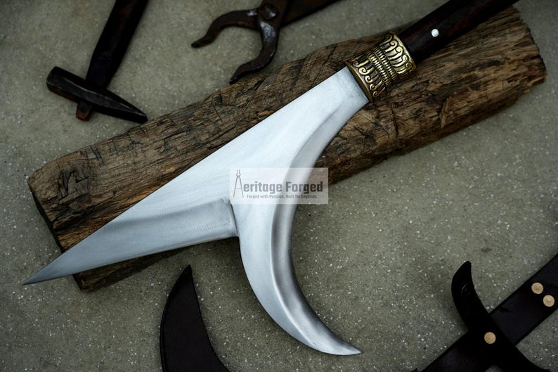 May include: A silver-colored curved blade with a wooden handle and a brass decorative piece. The blade is on a wooden surface. The text "Heritage Forged" is visible on the blade.