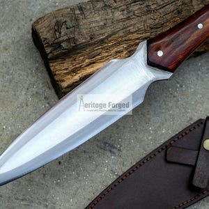 K&ouml;nnte beinhalten: Ein langes, scharfes, silbernes Messer mit einem Holzgriff. Das Messer hat eine braune Lederscheide mit einem Riemen und zwei Metallkn&ouml;pfen.