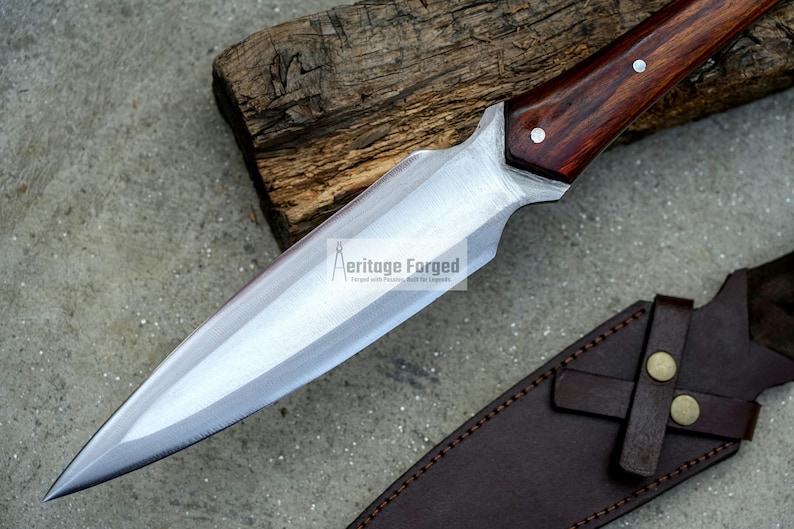 K&ouml;nnte beinhalten: Ein langes, scharfes, silbernes Messer mit einem Holzgriff. Das Messer hat eine braune Lederscheide mit einem Riemen und zwei Metallkn&ouml;pfen.