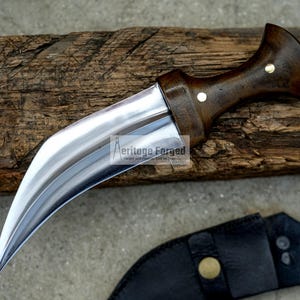May include: A curved, silver-coloured knife with a wooden handle. The handle has a dark brown finish and two brass accents. The knife is resting on a piece of weathered wood.