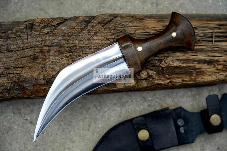 May include: A curved, silver-coloured knife with a wooden handle. The handle has a dark brown finish and two brass accents. The knife is resting on a piece of weathered wood.