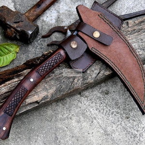 May include: A handcrafted, curved blade knife with a dark wood handle and a brown leather sheath. The handle features a diamond pattern and is accented with white rivets. The knife is resting on a weathered wooden surface, with a hammer and other tools in the background.