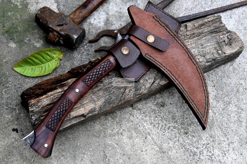 May include: A handcrafted, curved blade knife with a dark wood handle and a brown leather sheath. The handle features a diamond pattern and is accented with white rivets. The knife is resting on a weathered wooden surface, with a hammer and other tools in the background.