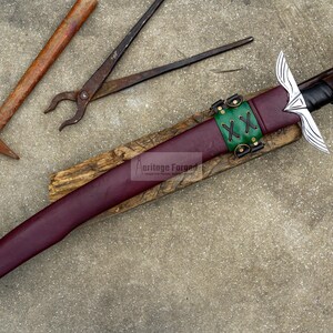 May include: A handcrafted sword with a burgundy leather sheath, green leather accents, and a silver blade. The sword rests on a wooden base, with a hammer and pliers in the background. The text "Heritage Forged" is visible on the wooden base.