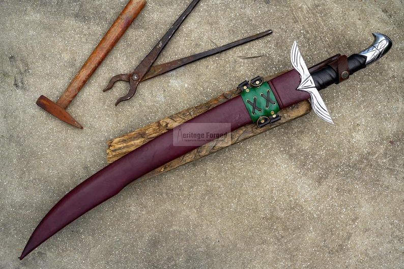 May include: A handcrafted sword with a burgundy leather sheath, green leather accents, and a silver blade. The sword rests on a wooden base, with a hammer and pliers in the background. The text "Heritage Forged" is visible on the wooden base.