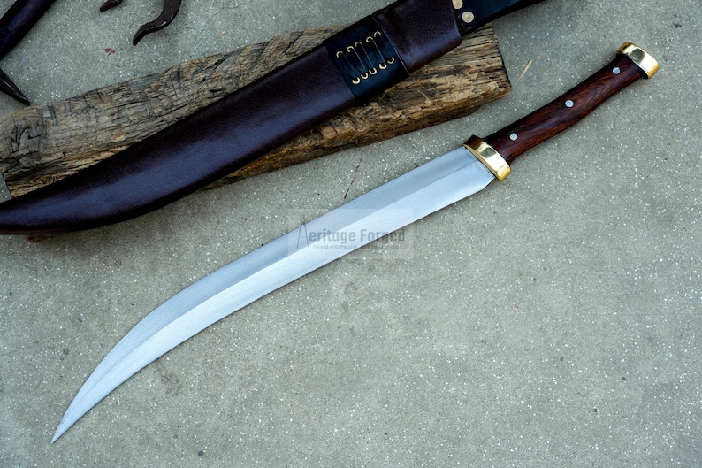May include: A curved, silver-colored knife with a wooden handle and brass accents. The knife is resting on a wooden surface and has a leather sheath.