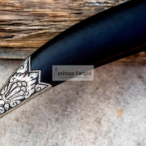 May include: Close-up of the ornate silver tip of a black leather-wrapped dagger. The tip features intricate floral designs and is pointed.