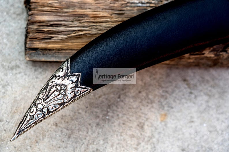 May include: Close-up of the ornate silver tip of a black leather-wrapped dagger. The tip features intricate floral designs and is pointed.