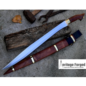 May include: A long, silver blade with a wooden handle and a brown leather sheath. The blade is sharp and pointed, and the handle is decorated with brass accents. The sheath has a brass accent at the top and a black leather strap.