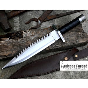 Pode incluir: Uma faca de prata com cabo preto e l&acirc;mina serrilhada. A faca est&aacute; apoiada em uma bainha de couro marrom. O texto "Heritage Forged" &eacute; vis&iacute;vel no canto inferior direito da imagem. Forjado com paix&atilde;o, constru&iacute;do para lendas.