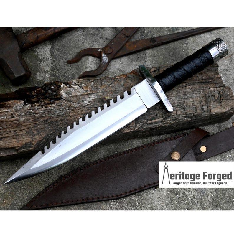 Pode incluir: Uma faca de prata com cabo preto e l&acirc;mina serrilhada. A faca est&aacute; apoiada em uma bainha de couro marrom. O texto "Heritage Forged" &eacute; vis&iacute;vel no canto inferior direito da imagem. Forjado com paix&atilde;o, constru&iacute;do para lendas.