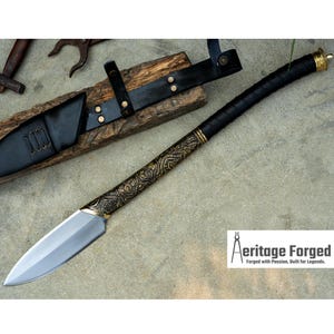 May include: A long, thin, silver blade with a black leather handle and gold accents. The blade has a decorative pattern etched into it. The handle is wrapped in black leather and has a gold pommel. The blade is resting on a wooden surface with a leather sheath.