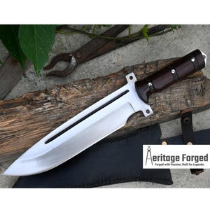 May include: A large, silver, fixed blade knife with a brown wooden handle. The knife has a black leather sheath and a full tang. The blade is sharp and has a slight curve. The handle is decorated with rivets. The knife is resting on a piece of wood.