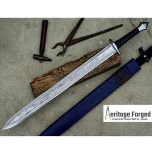 May include: A long, silver sword with an ornate blade design, black handle, and a blue sheath. The sword rests on a wooden plank with blacksmith tools nearby. The text "Aeritage Forged" is visible.