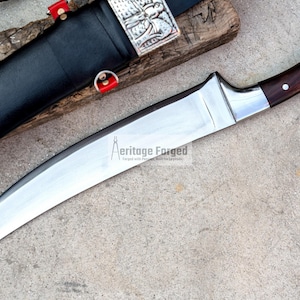 May include: A curved, silver-colored knife with a brown wooden handle. The handle has a decorative silver ring and a silver cap. The knife is on a gray background. The text "Heritage Forged" is printed on the blade.