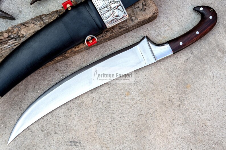 May include: A curved, silver-colored knife with a brown wooden handle. The handle has a decorative silver ring and a silver cap. The knife is on a gray background. The text "Heritage Forged" is printed on the blade.