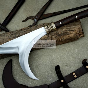 May include: A curved, silver-colored blade with a brown wooden handle and brass accents. The blade is attached to a wooden log. The text "Heritage Forged" is visible on the log.