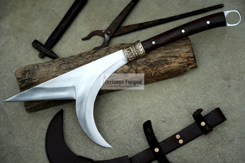 May include: A curved, silver-colored blade with a brown wooden handle and brass accents. The blade is attached to a wooden log. The text "Heritage Forged" is visible on the log.