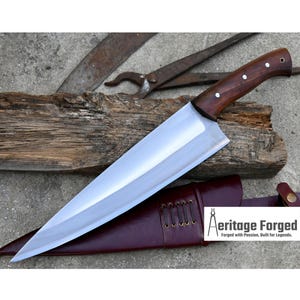 May include: A long, sharp, silver chef's knife with a wooden handle. The knife is resting on a piece of wood and a brown leather sheath. The text "Heritage Forged" is visible on the sheath. "Forged with Passion, Built for Legends."