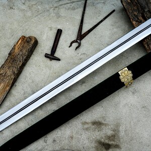 May include: A long, silver sword with a black handle and a decorative gold hilt, resting on a concrete surface. The sword is accompanied by a black sheath with gold accents, a small hammer, pliers, and a piece of wood.