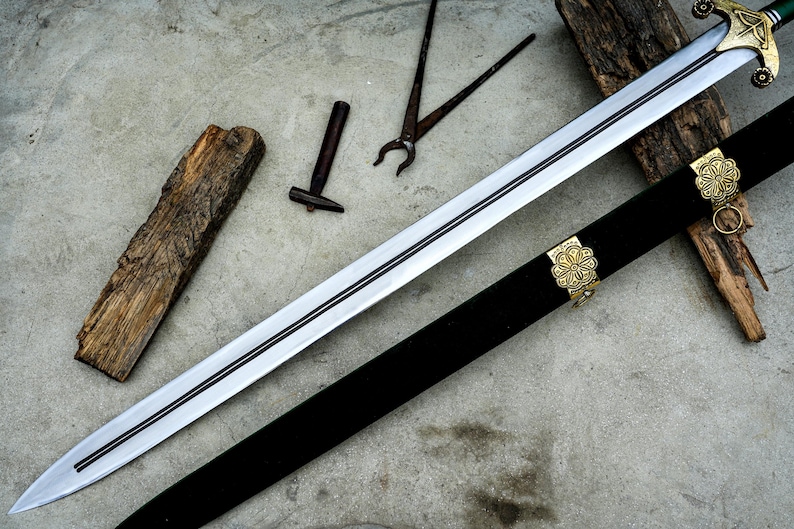 May include: A long, silver sword with a black handle and a decorative gold hilt, resting on a concrete surface. The sword is accompanied by a black sheath with gold accents, a small hammer, pliers, and a piece of wood.
