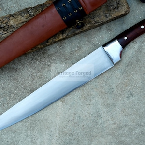 May include: A long, silver knife with a brown wooden handle. The knife has a sharp point and a curved blade. The knife is on a wooden surface with a brown leather sheath.