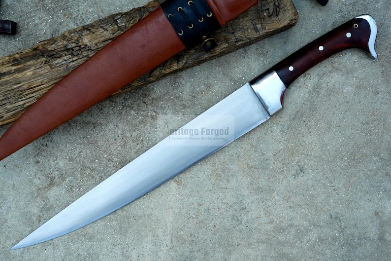 May include: A long, silver knife with a brown wooden handle. The knife has a sharp point and a curved blade. The knife is on a wooden surface with a brown leather sheath.