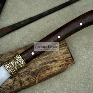 May include: A brown wooden handled knife with a silver blade and a gold decorative piece at the base of the handle. The knife is resting on a piece of wood. The text "Heritage Forged" is visible on the handle.