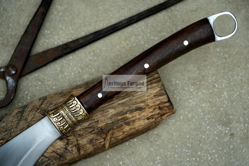 May include: A brown wooden handled knife with a silver blade and a gold decorative piece at the base of the handle. The knife is resting on a piece of wood. The text "Heritage Forged" is visible on the handle.