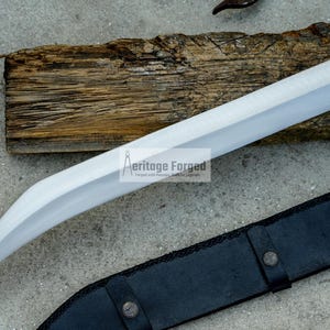 May include: A silver blade with a curved handle and a black leather sheath. The blade is sharp and has a polished finish. The sheath has a decorative design and is secured with a metal buckle. The text "Heritage Forged" is visible on the blade.