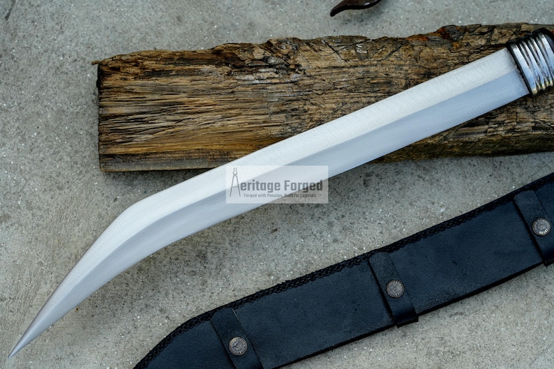 May include: A silver blade with a curved handle and a black leather sheath. The blade is sharp and has a polished finish. The sheath has a decorative design and is secured with a metal buckle. The text "Heritage Forged" is visible on the blade.