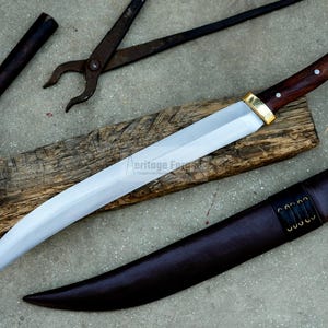 May include: A curved, silver-colored knife with a brown wooden handle and a gold-colored metal guard. The knife is resting on a piece of wood and has a brown leather sheath.