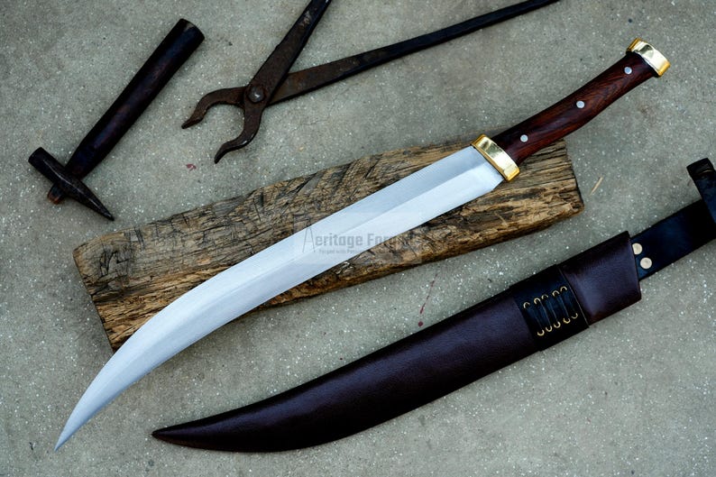 May include: A curved, silver-colored knife with a brown wooden handle and a gold-colored metal guard. The knife is resting on a piece of wood and has a brown leather sheath.