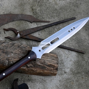 May include: A silver spear with a dark wood handle. The spear has a long, pointed blade with a series of holes along the top edge. The spear is resting on a piece of wood.