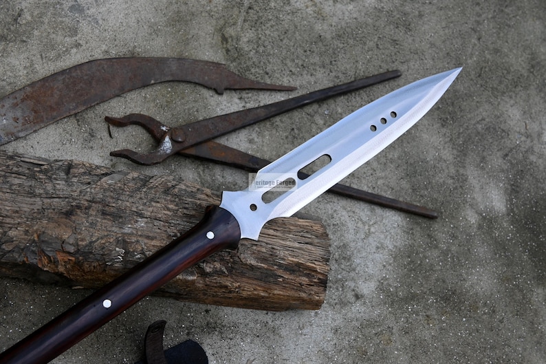 May include: A silver spear with a dark wood handle. The spear has a long, pointed blade with a series of holes along the top edge. The spear is resting on a piece of wood.