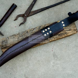 May include: A brown leather sheath with a black leather strap and brass accents. The sheath is wrapped around a knife with a wooden handle. The sheath is sitting on a piece of wood.