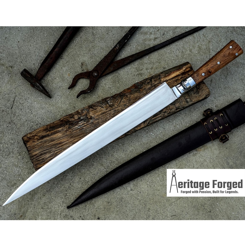 May include: A long, silver blade with a wooden handle. The blade is sharp and pointed, and the handle is made of dark wood with a brown leather sheath. The blade is resting on a piece of wood, and there are two metal tools in the background. The text "Heritage Forged" is visible in the lower right corner of the image. "Forged with Passion, Built for Legends."