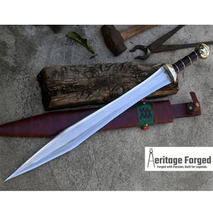May include: A polished steel sword with a brown leather handle and a gold-colored pommel. The sword rests on a brown leather sheath with green stitching. The image includes the text "Aeritage Forged: Forged with Passion, Built for Legends."