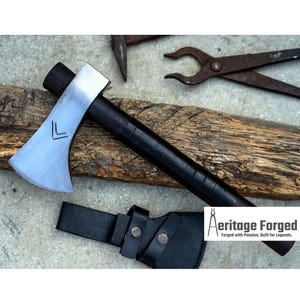 May include: A black-handled tomahawk with a silver blade and a leather sheath. The blade has a small, etched design. The tomahawk is resting on a piece of wood. The text "Heritage Forged" is visible in the lower right corner of the image. "Forged with Passion, Built for Legends."