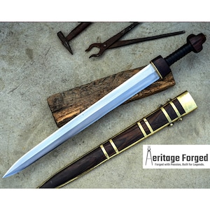 Könnte beinhalten: Ein silbernes Schwert mit einem Holzgriff und einer braunen Lederscheide mit goldenen Akzenten. Das Schwert liegt auf einem Holzblock und die Scheide liegt daneben. Der Text "Heritage Forged" ist in der unteren rechten Ecke des Bildes sichtbar. Geschmiedet mit Leidenschaft, gebaut für Legenden.