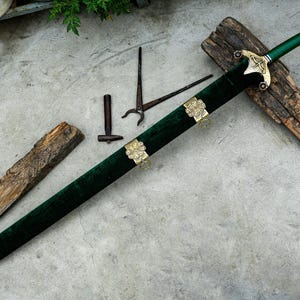 May include: A decorative sword with a green velvet scabbard and handle, featuring gold-colored accents. The sword rests on a wooden surface, with additional small metal tools nearby. The blade is not visible.
