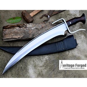 May include: A curved, silver-coloured sword with a brown wooden handle and a black leather scabbard. The sword has a decorative design on the blade and a small metal ring near the handle. The sword is lying on a grey surface with a wooden plank and a hammer in the background. The text "Heritage Forged" is visible in the lower right corner of the image.