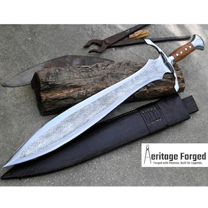 May include: A silver sword with a brown leather scabbard and a wooden handle. The sword has a decorative pattern etched into the blade. The sword is lying on a wooden surface with a metal tool in the background. The text "Heritage Forged" is visible in the lower right corner of the image. "Forged with Passion, Built for Legends."