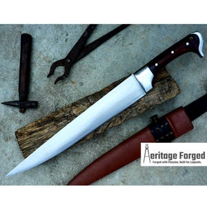 May include: A long, sharp, silver knife with a brown wooden handle. The knife is resting on a piece of wood and has a leather sheath. The text "Heritage Forged" is visible in the lower right corner of the image.  Forged with Passion, Built for Legends.