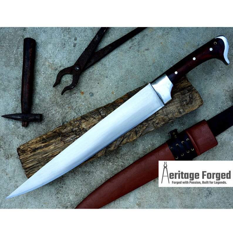 May include: A long, sharp, silver knife with a brown wooden handle. The knife is resting on a piece of wood and has a leather sheath. The text "Heritage Forged" is visible in the lower right corner of the image.  Forged with Passion, Built for Legends.