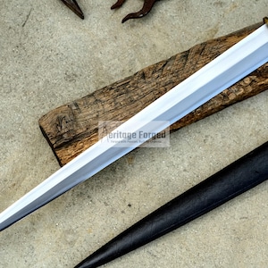 May include: A silver and gold dagger with a wooden handle rests on a wooden plank. A black leather sheath is positioned to the right of the dagger. The dagger has a sharp, pointed blade and a gold-colored guard.